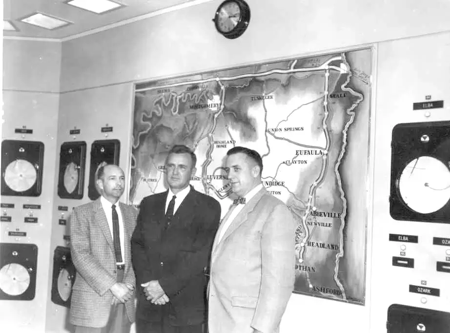 Three men in suits stand in a gas control room in front of a large wall map and monitoring gauges, circa mid-20th century.