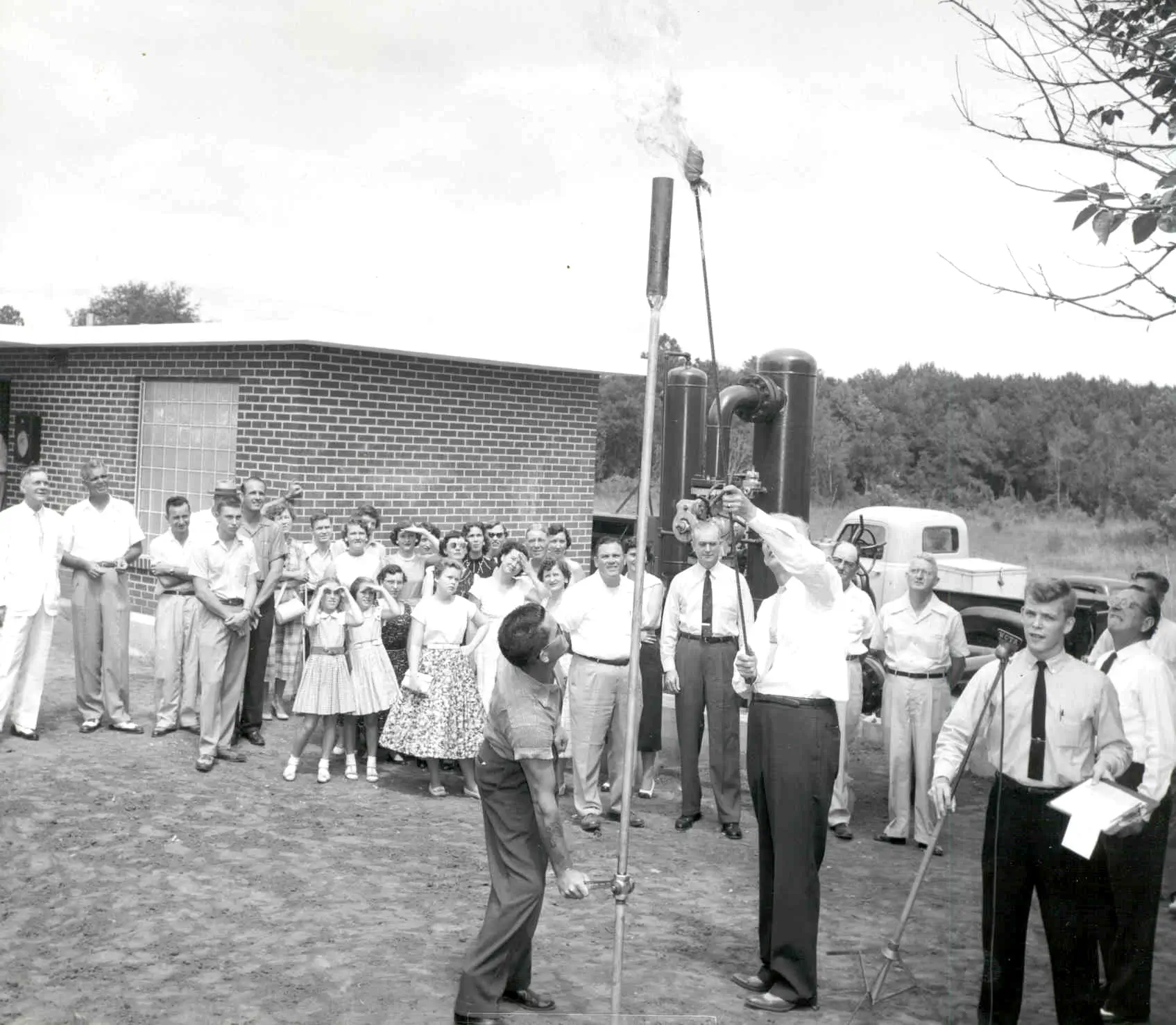 Historic photo of a crowd gathered as officials light a natural gas flare during a community ceremony, with men, women, and children watching outside a brick building.