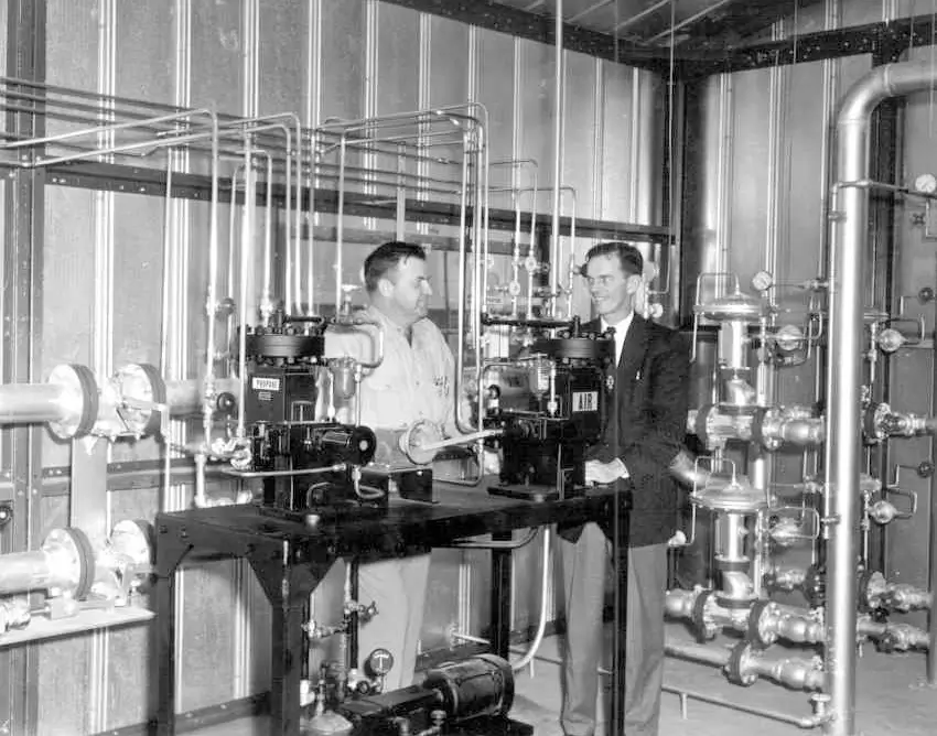 Two men, one in work clothes and one in a suit, stand inside a valve station surrounded by metal pipes and industrial equipment, mid-20th century.