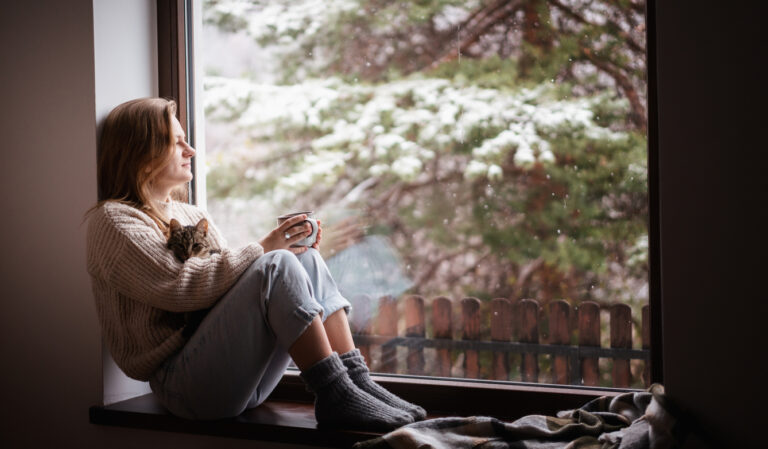 Young cheerful girl sitting at home on the windowsill in a warm sweater playing with a gray cat on a winter day